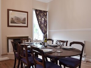 A dining room with a round table set for six, flanked by wooden chairs with blue cushions. A framed landscape painting adorns the wall above the fireplace.
