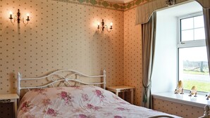 Una habitación con cama, dos lámparas fijadas a la pared, una ventana con cortinas y una mesita de noche.
