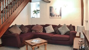 Een woonkamer met een houten trap, een rode bank met gekleurde kussens, een houten bijzettafeltje en een schilderij aan de muur.