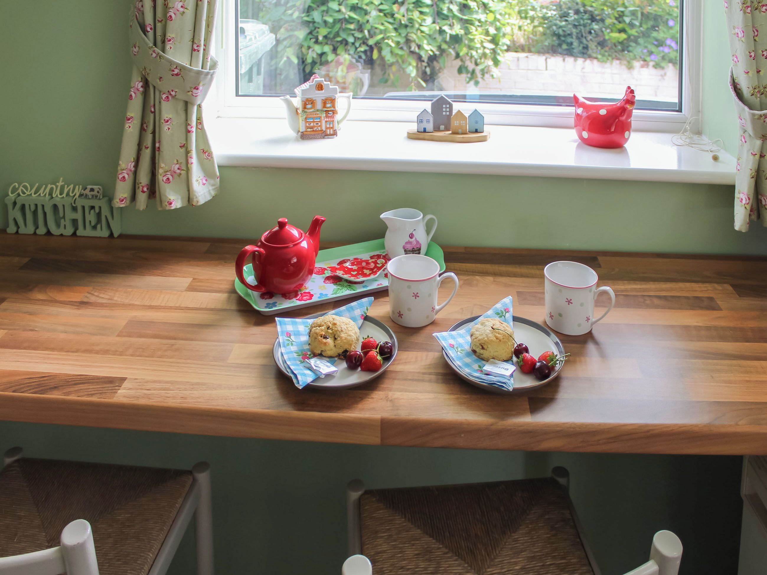 A cozy kitchen area with a wooden table set for tea, featuring a red teapot, white cups, and plates with biscuits and strawberries.