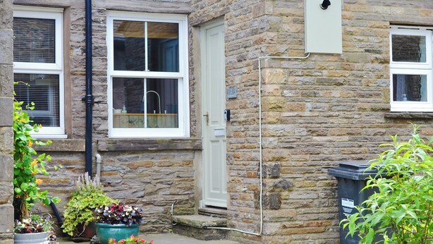 A stone house with a white door, two windows, and a small step leading to the entrance. There are potted plants on the step and a black trash bin to the right. - 2 bedroom accommodation in Burtersett, near Hawes (Burtersett, near Hawes)