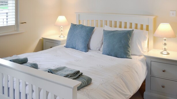 Una cama bien tendida con almohadas y una manta, dos lámparas de noche, una mesita de noche con un cajón y una ventana con persianas.