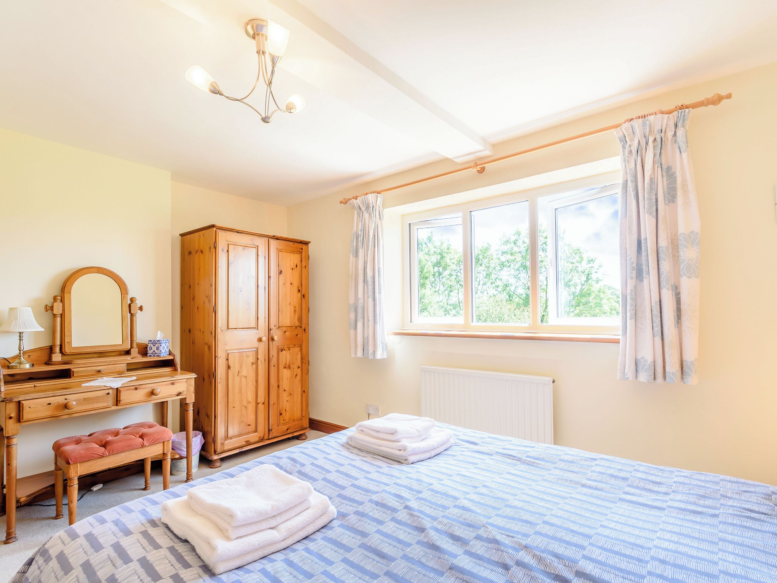 A bedroom with a bed, a wooden wardrobe, a dressing table with a mirror, and a window with curtains.