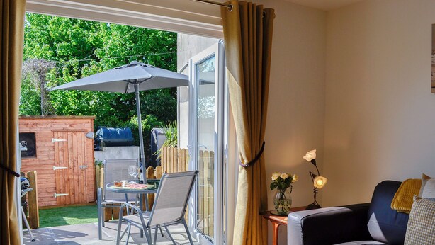 Ein Zimmer mit Glastür, die auf eine Terrasse führt, einem Sofa, einem kleinen Tisch mit einer Vase und einem Grill draußen.