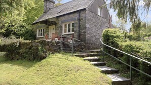 Una casa de piedra con techo inclinado, un pequeño porche con sillas y una escalera de piedra que conduce a la entrada.