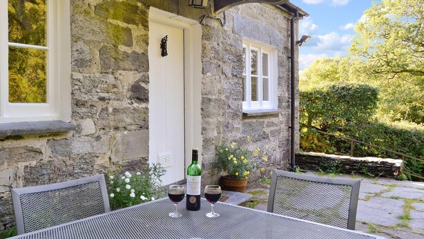 Un edificio de piedra con una puerta y ventanas blancas, una mesa con dos copas de vino tinto y una botella de vino.