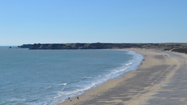 Ein weiter Sandstrand mit sanften Wellen, wolkenlosem Himmel und fernen Klippen.