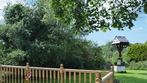 A wooden deck with a railing, surrounded by lush greenery and a birdhouse. - Hunter's Cottage - UK6239 (Y Ff&ocirc;r, near Pwllheli)