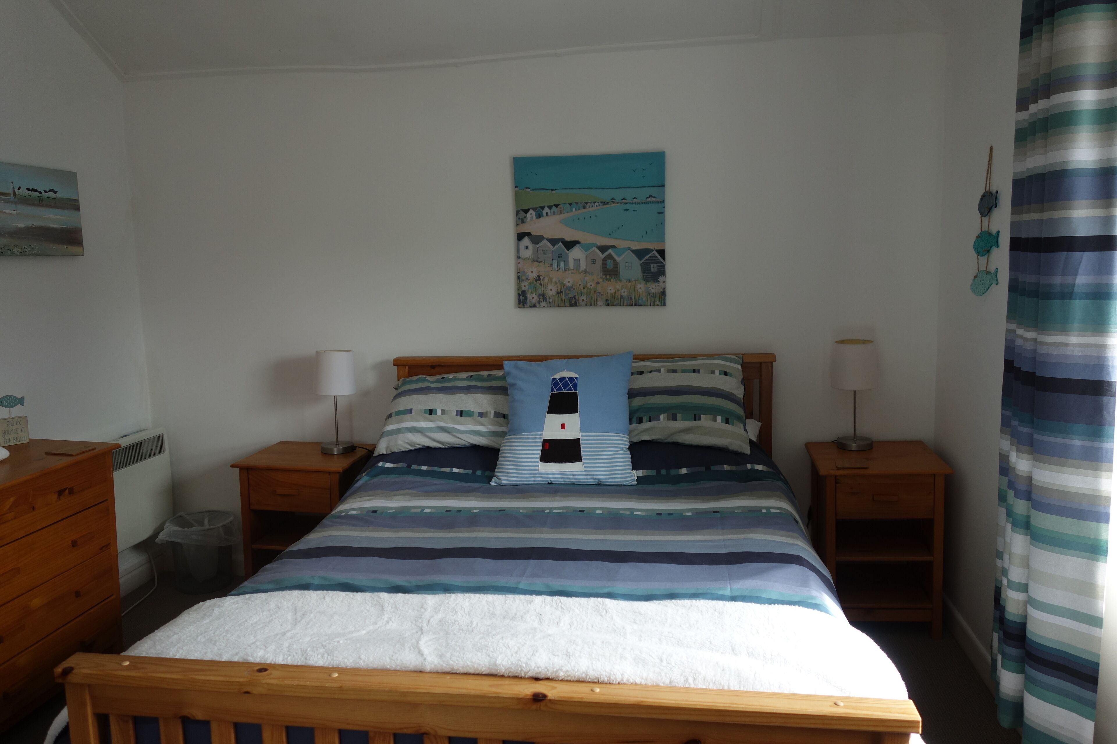A bedroom with a bed, two bedside tables, a wooden dresser, a painting on the wall, and a striped curtain.