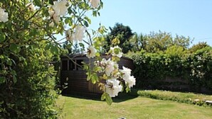 Un jardin avec un abri en bois, des fleurs blanches et un coin pique-nique avec une couverture à rayures.