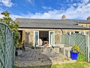 A stone house with a tiled roof, a garden with wicker furniture, and a blue planter.