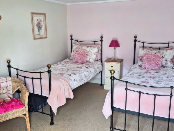 A bedroom with two twin beds, a nightstand, a lamp, a wicker chair, and a teddy bear.