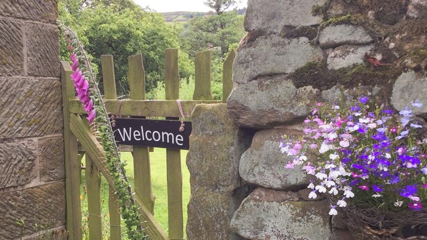 Una verja de madera con un cartel que dice 'Welcome', rodeada de follaje y un muro de piedra.