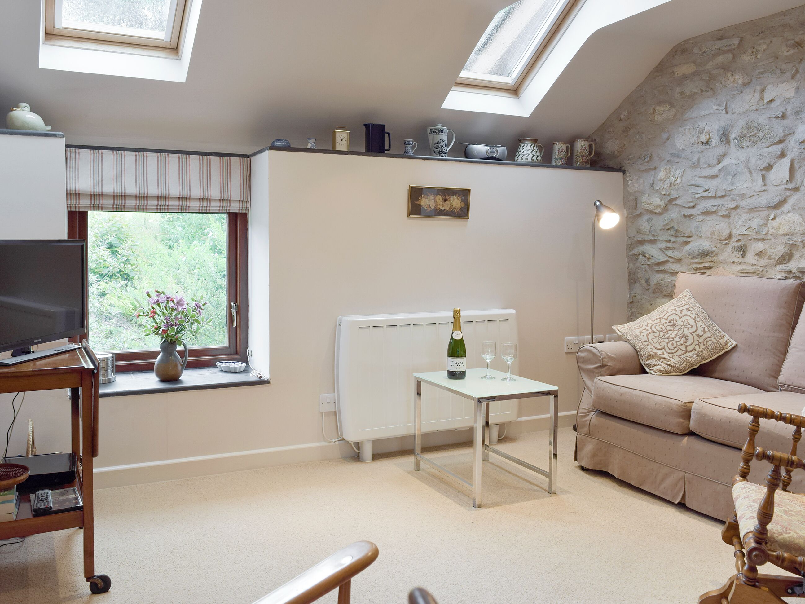 A cozy living room with a stone wall, a sofa, a TV, a small table with a bottle and glasses, and a window with a view of greenery.