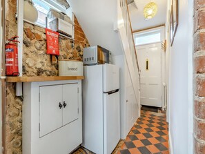 Un pasillo estrecho con pared de piedra, una manta ignífuga, un recipiente para pan, un refrigerador blanco y un suelo a cuadros.