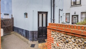 A narrow alleyway with a black door, a brick wall with a stone base, and a white building with a blue door and windows. - 3 bedroom accommodation in Cromer (Cromer)
