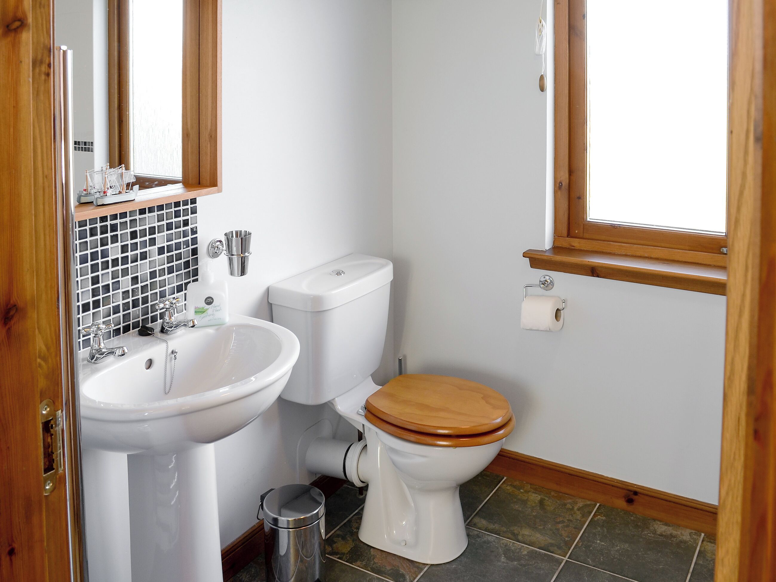 A bathroom with a white sink, toilet, and mirror. There is a window above the toilet and a trash can next to the sink.