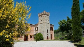 A historic building with a stone tower, surrounded by a gravel courtyard and lush greenery. - Bacco (Montefalco)