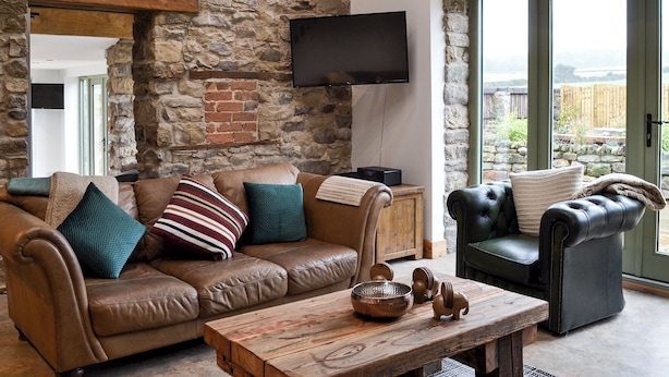 Un salon chaleureux avec un mur en pierre, des canapés en cuir, une table basse en bois et un téléviseur fixé au mur.