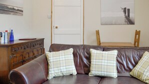 A living room with a brown leather sofa, two plaid cushions, a wooden side table, and a painting of a bridge on the wall.