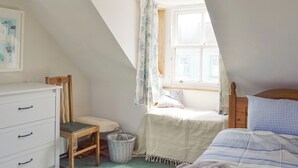 A bedroom with a sloped ceiling, a bed with a blue checkered pillow, a wooden chair, a white dresser, and a window with floral curtains.