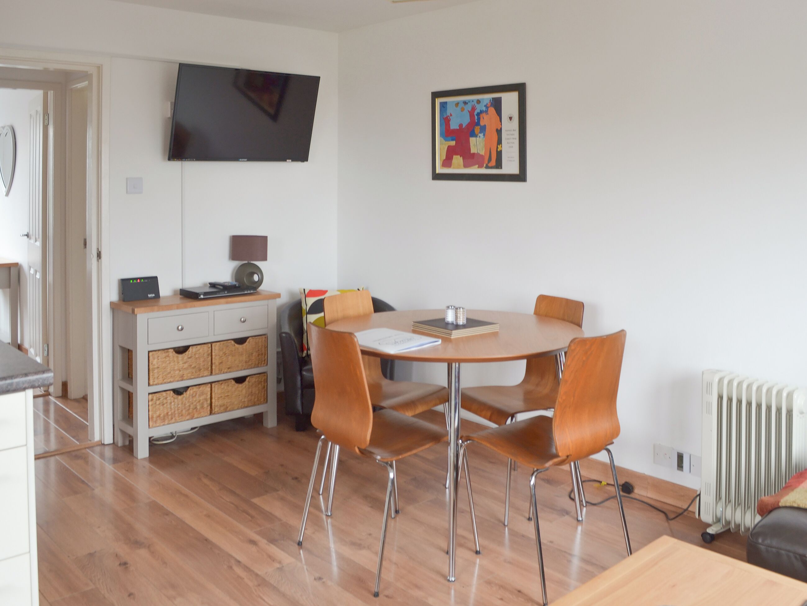 Et moderne spiseområde med et rundt bord, fire stole, et fjernsyn ophængt på væggen og et indrammet kunstværk over bordet.