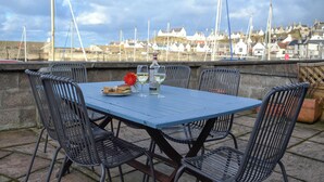 A table with chairs, a bottle, and a glass of white wine, set against a backdrop of a harbor with boats and buildings.