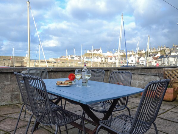 A table with chairs, a bottle, and a glass of white wine, set against a backdrop of a harbor with boats and buildings.