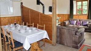 A dining area with a wooden table set for a meal, a staircase, and a living room with a brown leather sofa. - The Coach House - EXG (Upton, near Dulverton)