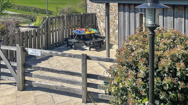 Un edificio de piedra con una valla de madera, una mesa de picnic con objetos de colores y un letrero que dice 'THE SMITHY'.