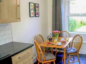A dining area with a wooden table, chairs, a coffee pot, a vase with flowers, and a window with a view of greenery.