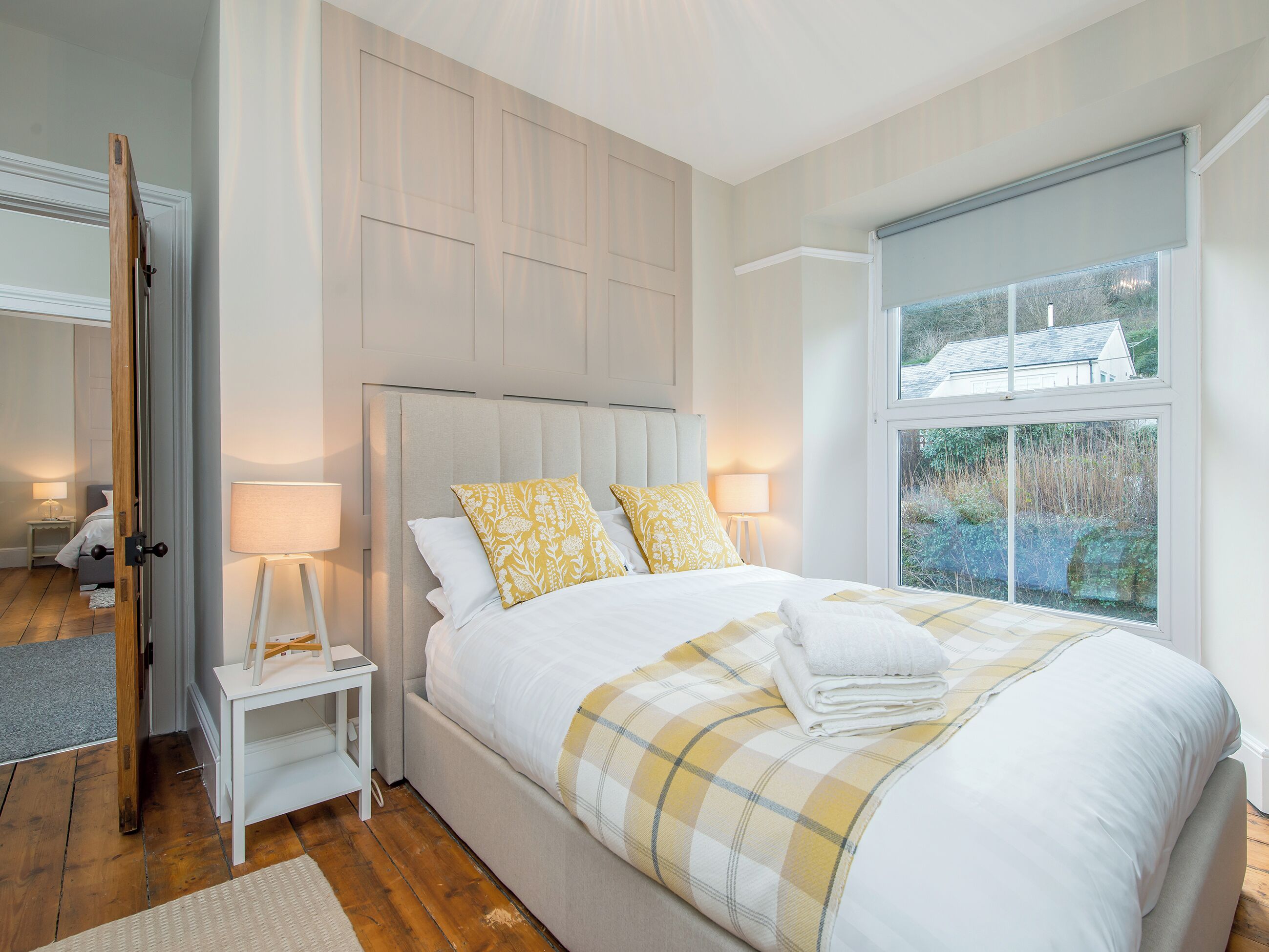 A bedroom with a large bed, two bedside tables with lamps, a window with a view of greenery, and a door leading to another room.