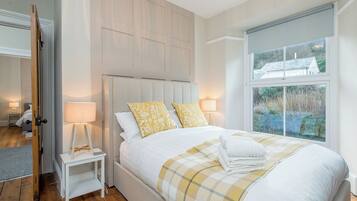 A bedroom with a large bed, two bedside tables with lamps, a window with a view of greenery, and a door leading to another room.