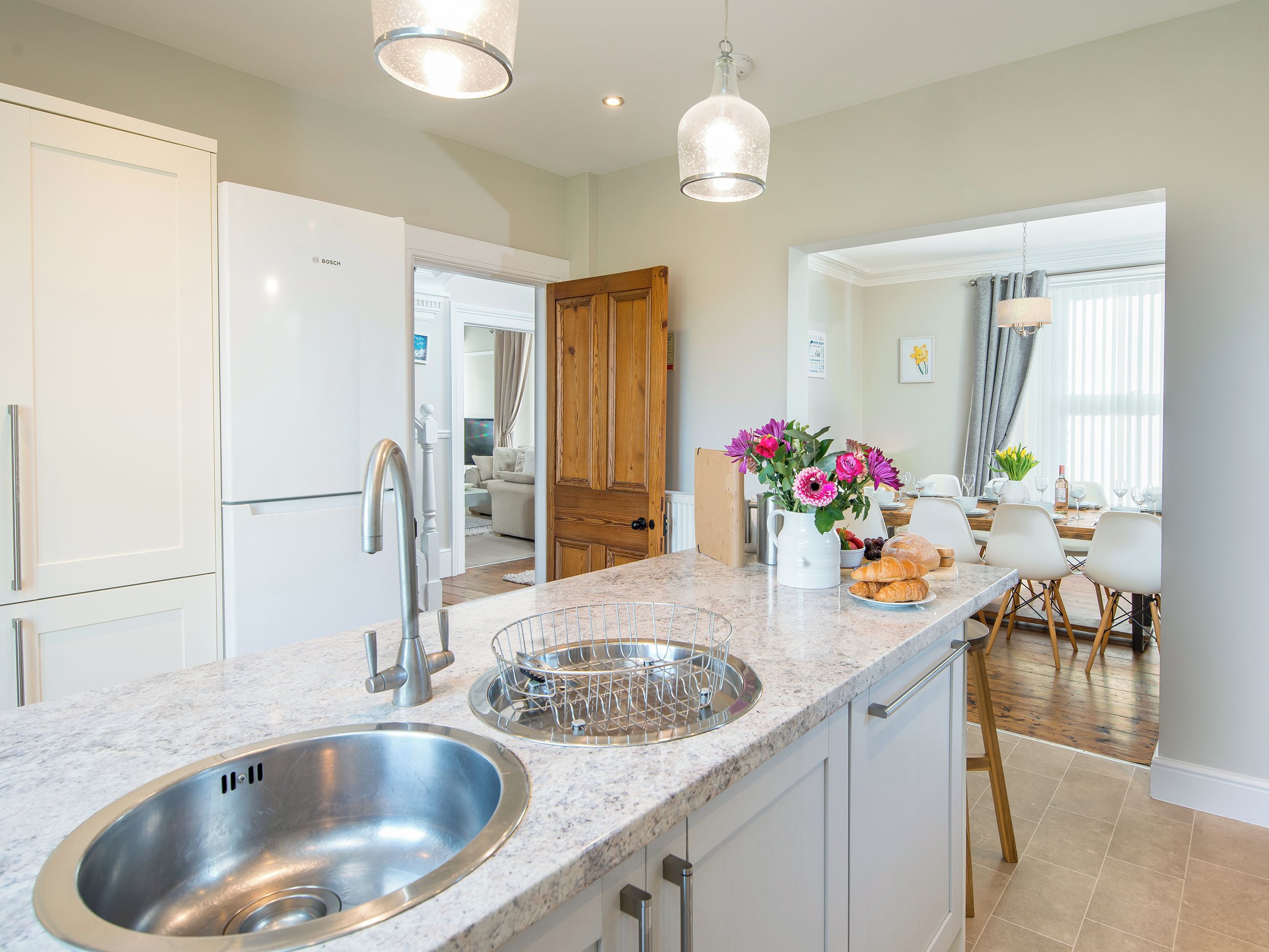 Una cocina moderna con un lavamanos de acero inoxidable, armarios blancos y encimera de mármol. Hay una puerta de madera que conduce a otra habitación y una zona de comedor con una mesa preparada para cuatro.