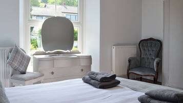Een slaapkamer met een bed, een dressoir, een stoel, een radiator en een raam met uitzicht op huizen.