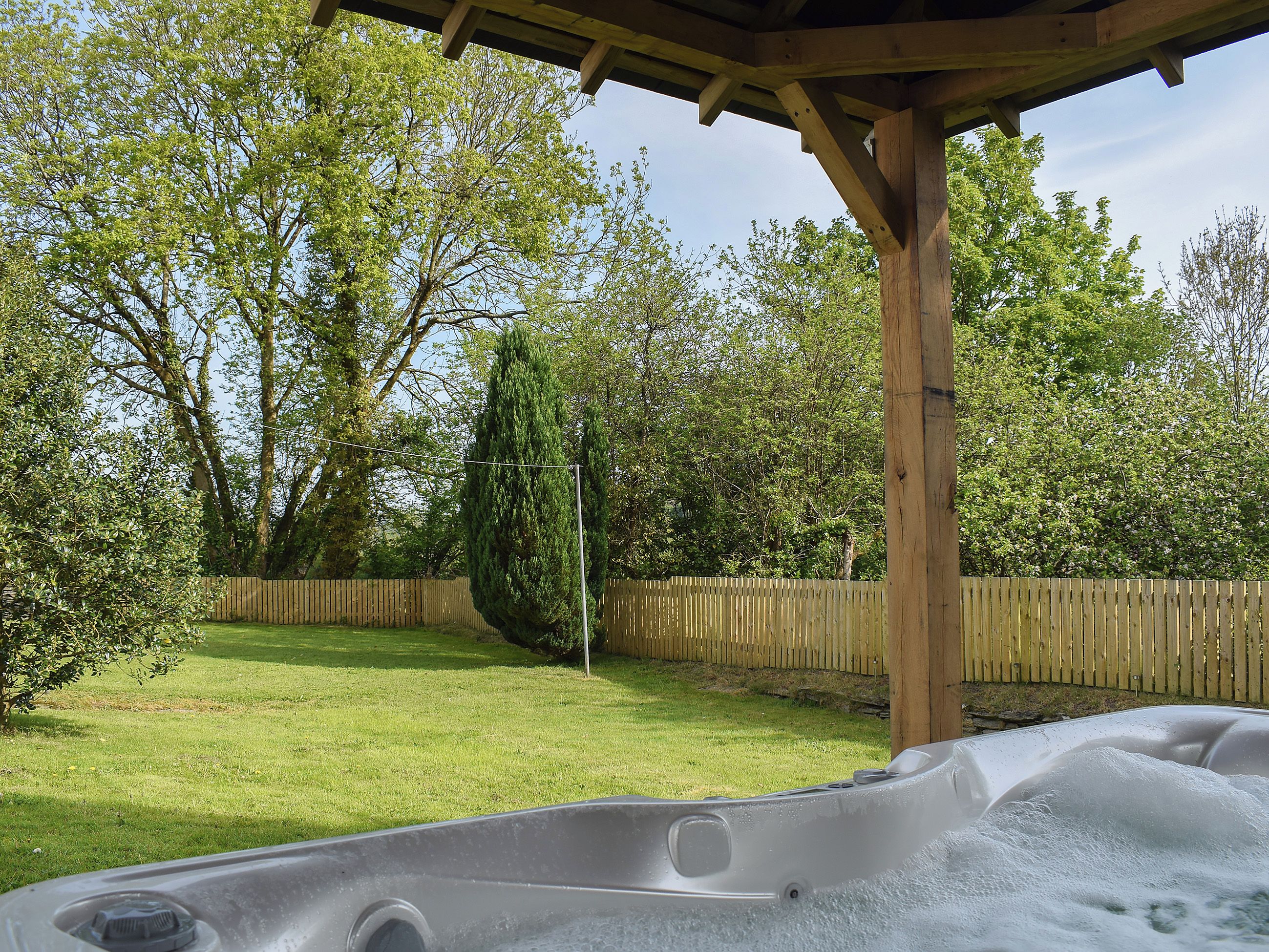 Un jacuzzi bajo una pérgola de madera en un patio trasero con un cerco de madera y árboles.
