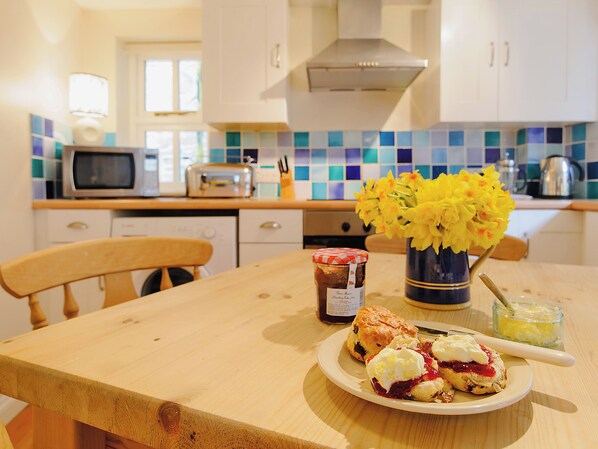A kitchen with a wooden table, a plate of scones, a jar of jam, and a vase of flowers.