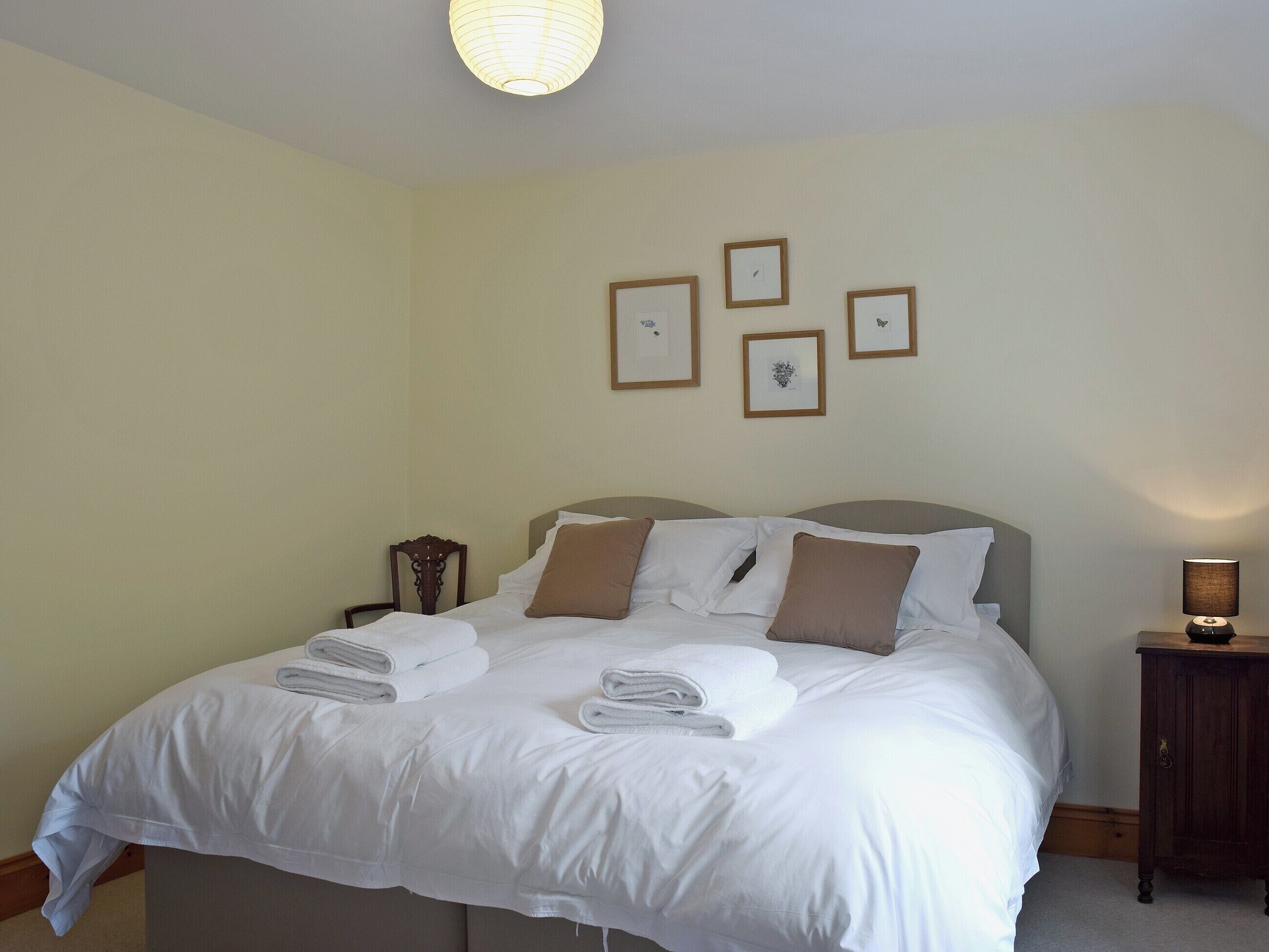 Una cama bien hecha con sábanas blancas y almohadas marrones, una mesita de noche con una lámpara, y cuadros enmarcados en la pared.