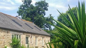 Un bâtiment en pierre avec un toit en pente, un petit jardin avec différentes plantes et un grand arbre en arrière-plan.