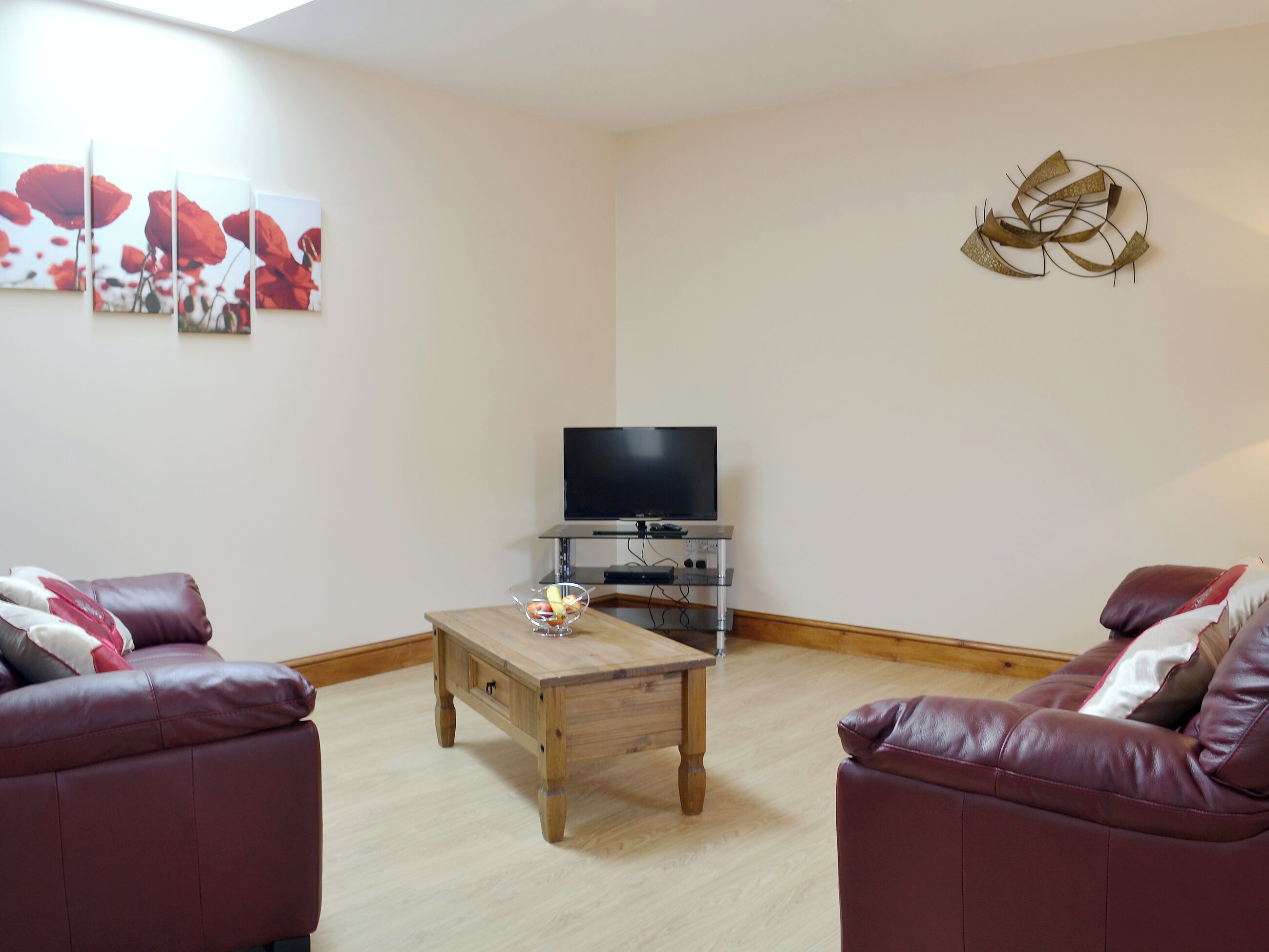 Ein Wohnzimmer mit einem Holztisch, zwei Ledersofas, einem Flachbildfernseher auf einem Standfuß und drei gerahmten Kunstwerken an der Wand.