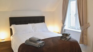 A neatly made bed with white linens and a brown blanket, a wooden nightstand with a lamp, and a window with curtains. - 1 bedroom accommodation in Whitby (Whitby)