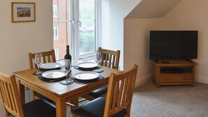 A dining area with a table set for four, a television on a stand, and a window with blinds. - 1 bedroom accommodation in Whitby (Whitby)