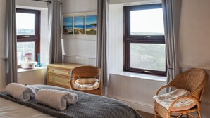 Ein Schlafzimmer mit einem Bett, einer Kommode, zwei Korbstühlen und einem Fenster mit einem Gemälde darüber.