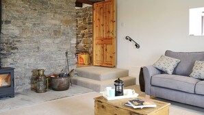 A cozy living room with a stone wall, a fireplace, a wooden door, a gray sofa, and a coffee table with books and a coffee pot. - 4 bedroom accommodation in Holy Island, near Berwick-upon-Tweed (Holy Island, near Berwick-upon-Tweed)