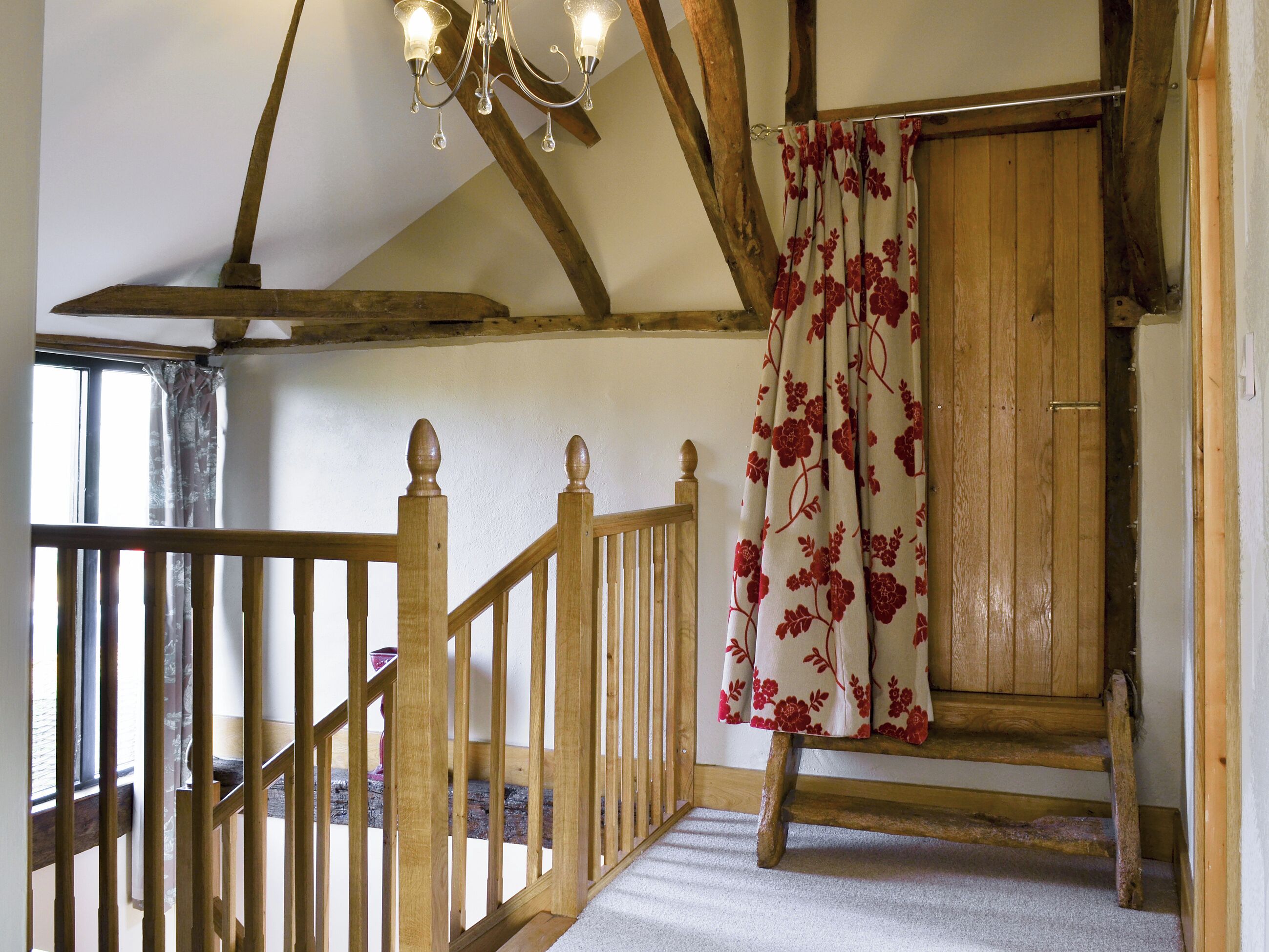 Un escalier en bois orné d’un lustre, une porte en bois avec des rideaux à motifs floraux et une vue sur l’extérieur.