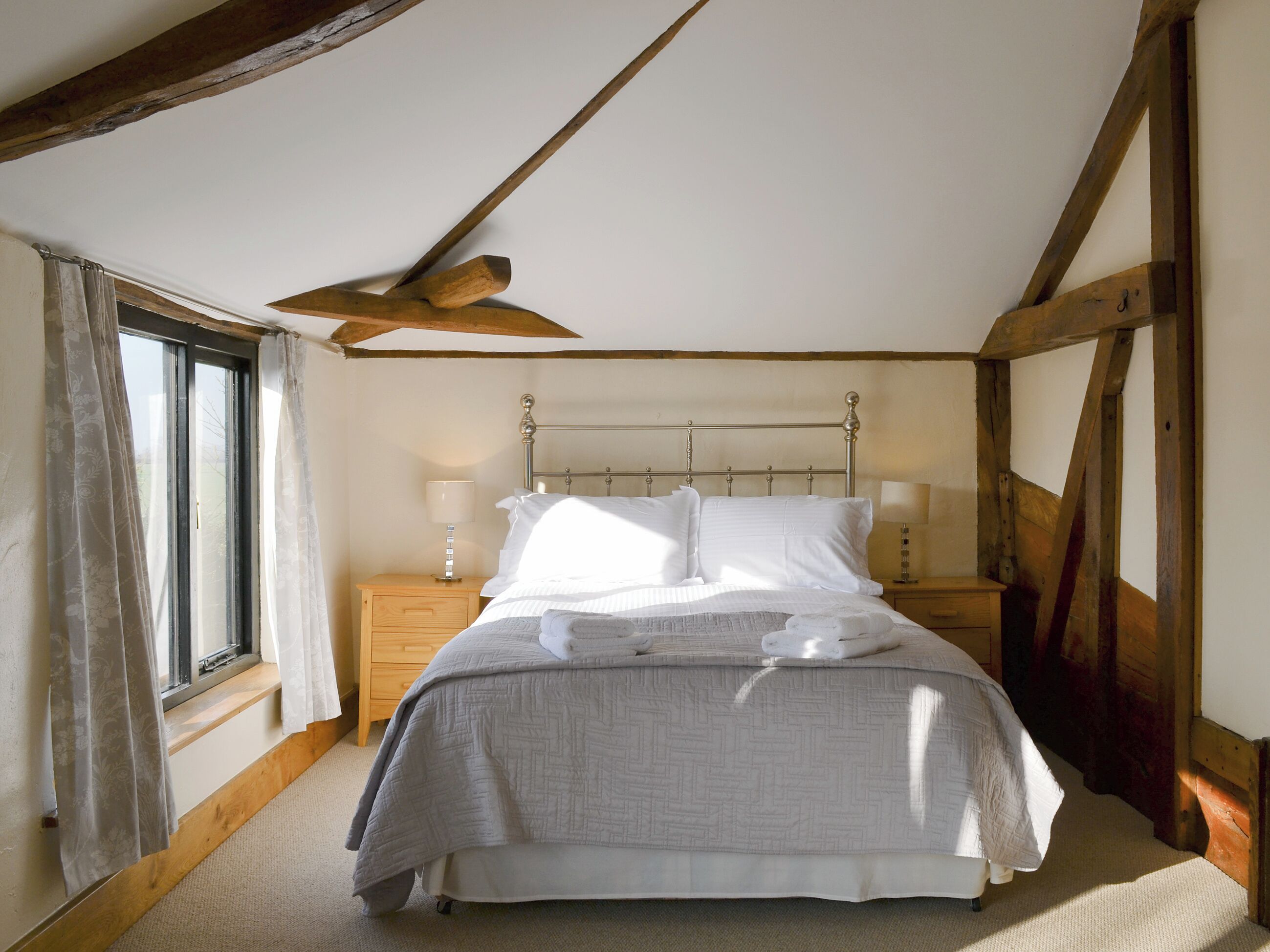 A bedroom with a bed, two bedside tables, a window with curtains, and wooden beams on the ceiling.