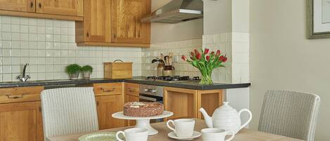 A kitchen with wooden cabinets, a tiled backsplash, a clock, a vase with flowers, and a dining table set with white cups and plates.