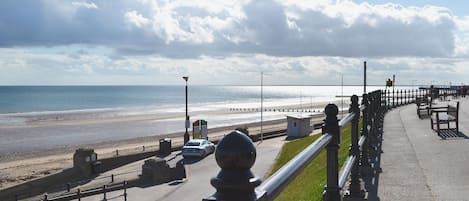 Une promenade en bord de mer, agrémentée d’une rambarde en métal, de bancs et offrant une vue dégagée sur la plage et la mer.