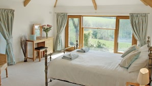 A bedroom with a wooden ceiling, a large window, a bed with pillows, a bedside table, and a desk with a vase of flowers. - 6 bedroom accommodation in Tregoodwell, Camelford (Tregoodwell, Camelford)