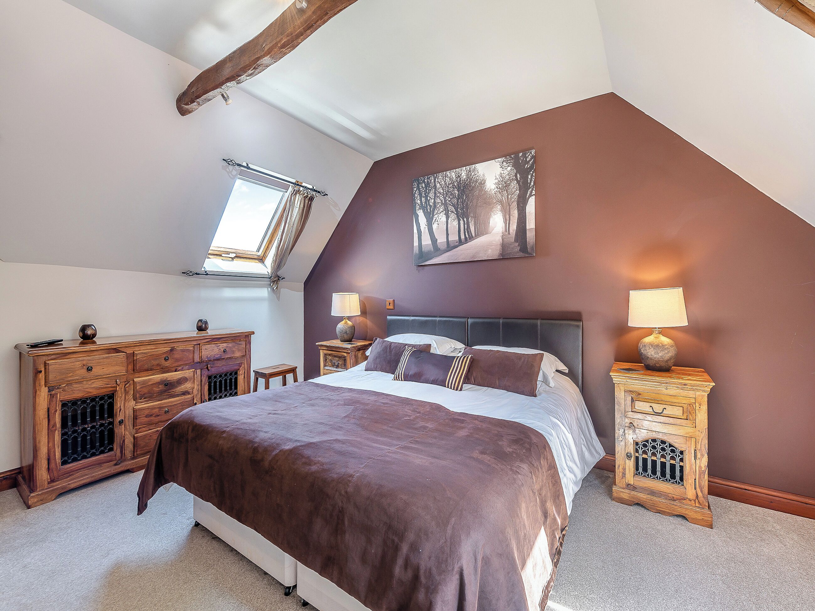 A bedroom with a sloped ceiling, a bed with a brown bedspread, two bedside tables with lamps, a wooden dresser, and a painting of trees on the wall.